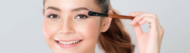 Closeup beautiful girl with flawless applying glamorous eye shadow makeup with eyeliner brush. Cosmetic facial painting process on lovely young woman with perfect clean skin in isolated background. - Фото, изображение