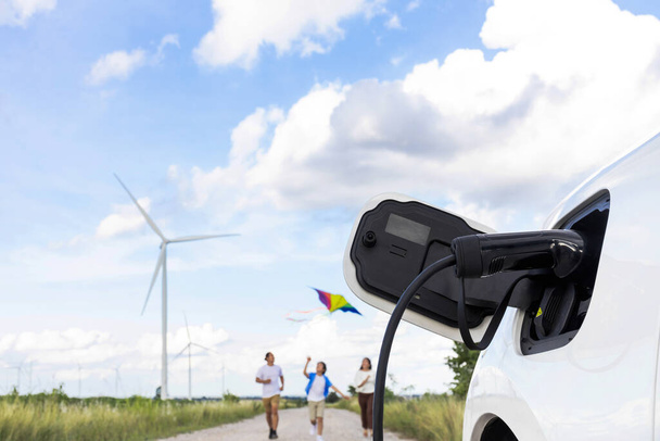 Concept of progressive happy family holding windmill toy and relax at wind farm with electric vehicle. Electric vehicle driven by clean renewable energy from wind turbine generator to charger station. - Zdjęcie, obraz