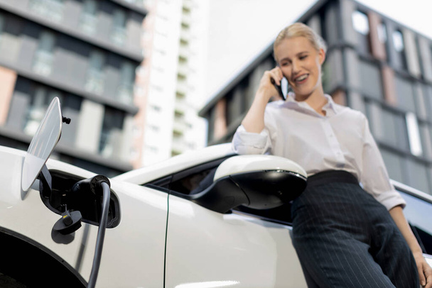 Focus charging-electric car with EV charger at charging station with blur businesswoman talking on phone with residential building in background as progressive lifestyle concept. - Fotografie, Obrázek