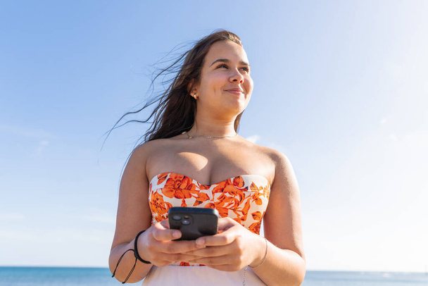 Un'adolescente usa il suo smartphone felice mentre si trova in riva al mare con il cielo limpido - Foto, immagini