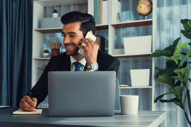 Трудолюбивый бизнесмен занят разговорами по телефону с клиентами, работая с ноутбуком в своем офисе, как концепция современного трудолюбивого офисного образа жизни с мобильным телефоном. Пылкий - Фото, изображение