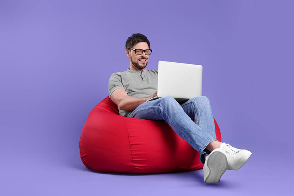 Hombre feliz con el ordenador portátil sentado en la silla del bolso de frijoles contra el fondo lila - Foto, imagen