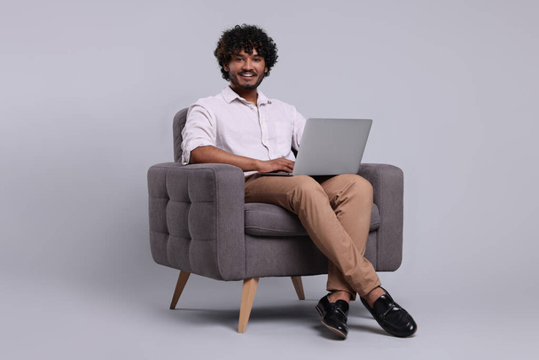 Smiling man with laptop sitting in armchair on light grey background, space for text - Foto, Imagem