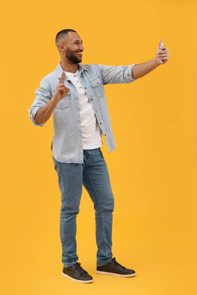 Smiling young man taking selfie with smartphone and showing peace sign on yellow background - Foto, afbeelding