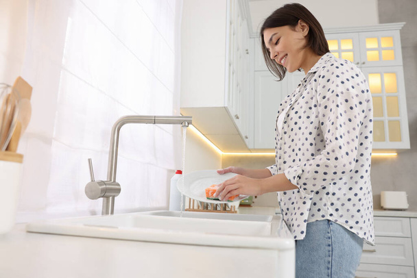 Happy young woman washing plate above sink in modern kitchen - Foto, afbeelding