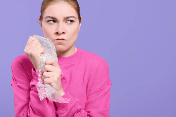 Angry woman popping bubble wrap on purple background, space for text. Stress relief - 写真・画像