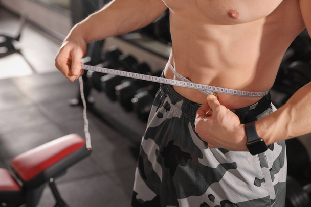 Athletic man measuring waist with tape in gym, closeup - Fotó, kép