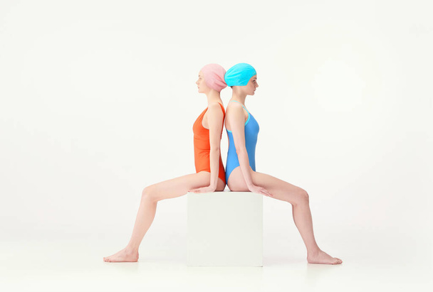 Two young girls, swimming athletes sitting in colorful retro swimsuits and swimming caps isolated over white background. Concept of retro style, sport, fashion, youth, vintage, competition - 写真・画像