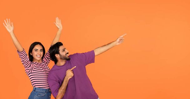 Szczęśliwa wesoła emocjonalnie tysiącletnia para Indian bawiąc się razem i pokazując puste miejsce na tekst lub reklamę na pomarańczowym tle studio, wskazując na świetną ofertę, web-banner - Zdjęcie, obraz