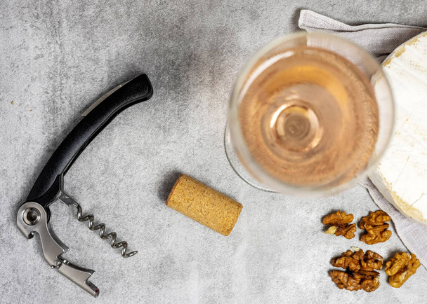 composition from rose wine bottle,glass, round cheese with one cut slice,walnuts,cork and corkscrew on kitchen striped towel or gray background tiles imitation.bottle upside down,flat lay front view - Fotó, kép