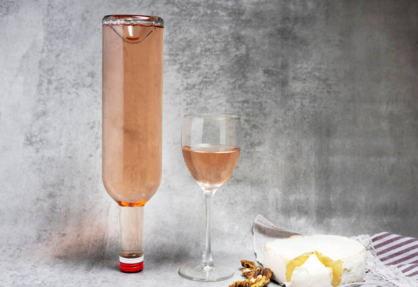 composition from rose wine bottle,glass, round cheese with one cut slice,walnuts,cork and corkscrew on kitchen striped towel or gray background tiles imitation.bottle upside down,flat lay front view - Photo, Image