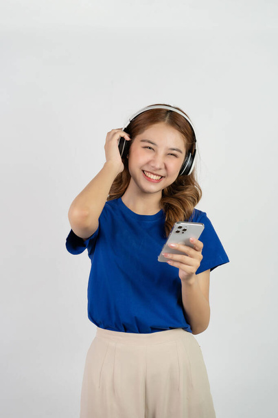 Adorable Asian girl holding smartphone and wearing headphones while listening to music with her blissful smile. standing against a white background - Foto, Bild