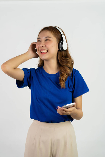 Urocza Azjatka trzymająca smartfona i nosząca słuchawki podczas słuchania muzyki z jej błogim uśmiechem. stojąc na białym tle - Zdjęcie, obraz