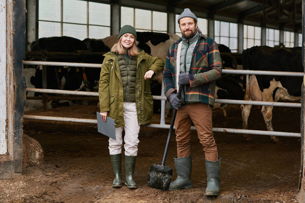 Portrait of farmers looking at camera standing in cowshed on dairy farm with cows in background - Foto, Bild