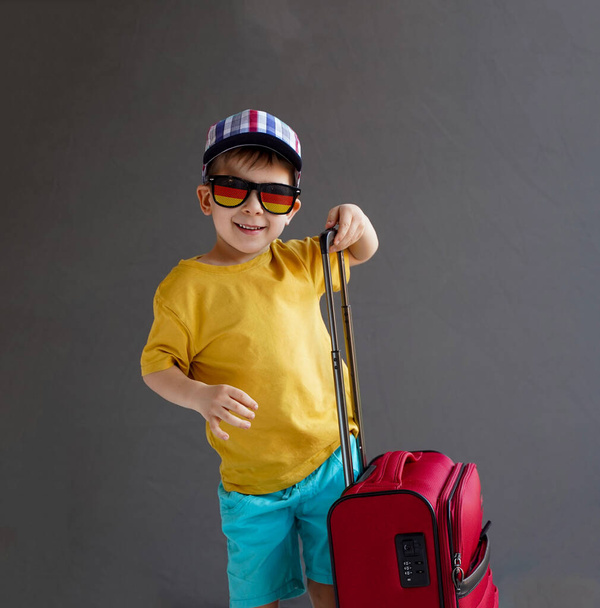 Un enfant en vêtements d'été et avec une valise sur fond gris. Photo du studio. Le concept des vacances d'été - Photo, image
