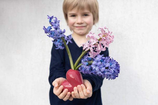 3 grote prachtige hyacint bloemen in hartvormige vaas in handen van kind op de voorgrond, gezicht van schattige lachende jongen in onscherp zicht. Moederdag, Verjaardag. Feestelijke stemming, aangename bloemenverrassing - Foto, afbeelding