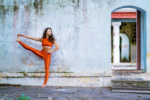 A young brunette woman in orange sportswear stretching her leg near an old building - Foto, Bild
