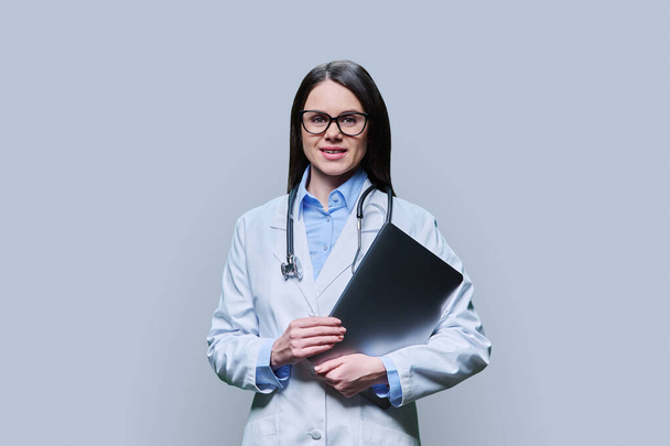 Young friendly female doctor with laptop computer on light gray studio background. Confident female in white coat with glasses looking at camera. Medical staff occupation health care science medicine - Fotó, kép