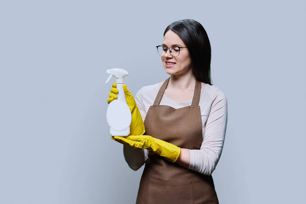 Young woman in gloves apron with organic detergent in hands looking at sprayer on light gray background. Home cleaning service, housekeeping cleanliness, natural eco organic detergents, ecology - Photo, Image