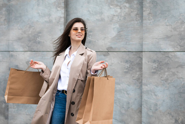 A girl in a casual clothes with shopping bags and lots of purchases near the shopping center smiling hands you her package, consumerism sale and people concept. - Photo, Image