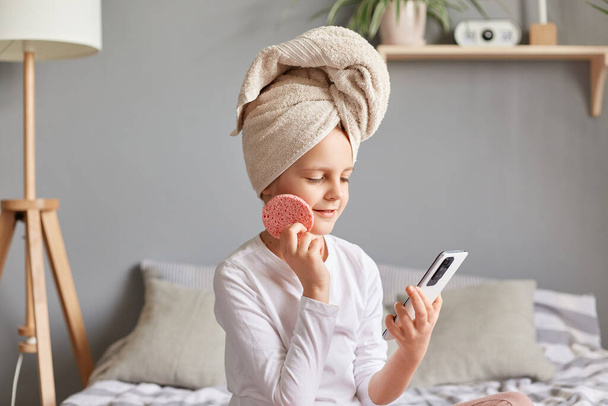 Cute charming little girl wearing white shirt and towel on head holding mobile phone and cosmetic sponge doing cosmetic procedures in the morning. - Valokuva, kuva