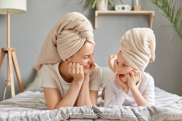 Delighted cute family resting after taking shower, mother and daughter laying in bed wearing towel posing in bedroom at home, looking at each other and talking. - Foto, immagini