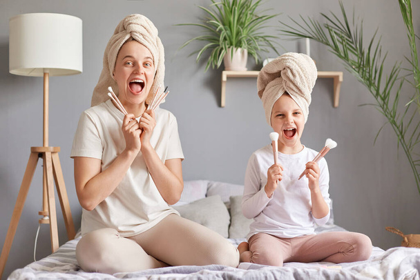Joyful screaming mother and daughter in towels on bed holding powder brushes yelling having fun at home having spa beauty day together doing each other makeup. - Foto, afbeelding