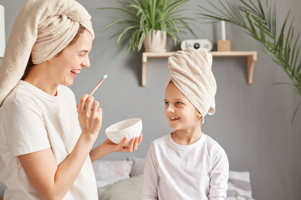 Portrait of mother and little daughter with facial masks on bed at home doing cosmetic procedures together beauty weekend having fun smiling. - Фото, зображення