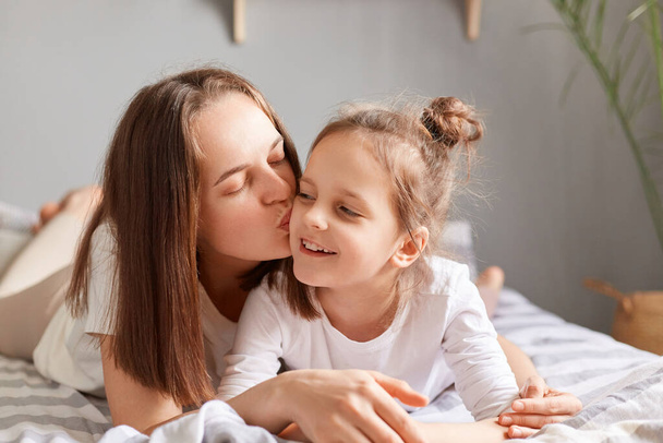 Portrait of smiling cute little girl lying in bed with mother, woman kissing her daughter with love, celebrating mother's day, enjoying lazy weekend, hugging expressing gentle emotions. - Фото, изображение