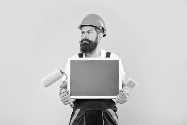 man in helmet and work clothes hold paint roller brush and blackboard with copy space on yellow background. - Foto, immagini