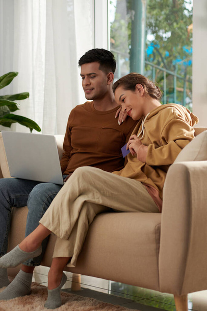Молодая пара влюбленных сидит дома на диване и платит за стриминговый сервис, чтобы смотреть любимое шоу онлайн - Фото, изображение