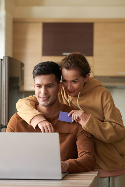 Young woman with credit card hugging boyfriend from behind when they are choosing food to order online - Photo, image
