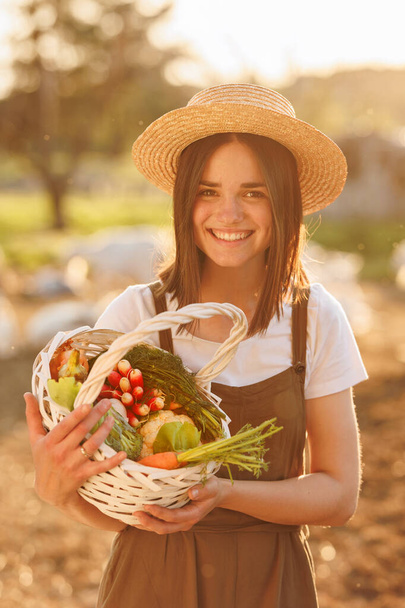 Młoda biała kobieta rolnik w słomkowym kapeluszu trzymając koszyk ze świeżymi organicznymi warzywami o zachodzie słońca. Wegetariańskie, surowe zdrowe jedzenie. Zdrowy styl życia. Gospodarstwo ekologiczne i działalność rolnicza. - Zdjęcie, obraz