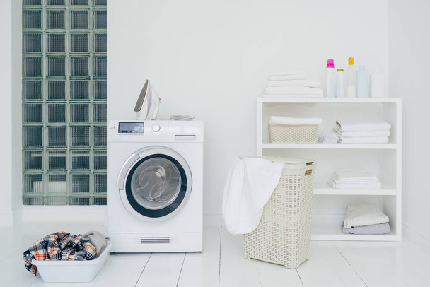 Laundry room with washing machine, dirty clothes in basket, iron and little shelf with neatly folded linen. Domestic room interior. Washing concept - Photo, Image