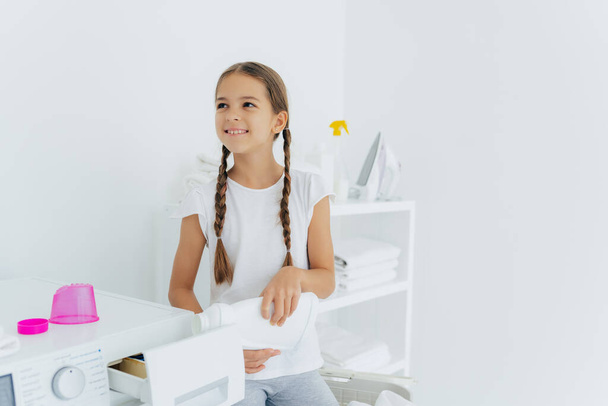 Little happy girl has two pigtails fills in washing machine with liquid detergent, pours into tray of washer, stands in laundry room with white walls, helps mother with washing, wears white t shirt - Foto, Bild