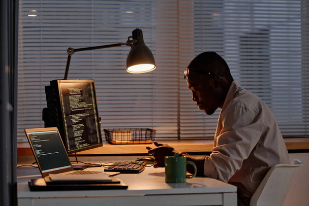 Afrikkalainen amerikkalainen IT-asiantuntija käyttää älypuhelinta työskennellessään koodien kanssa tietokoneella työpaikallaan myöhään yöllä - Valokuva, kuva