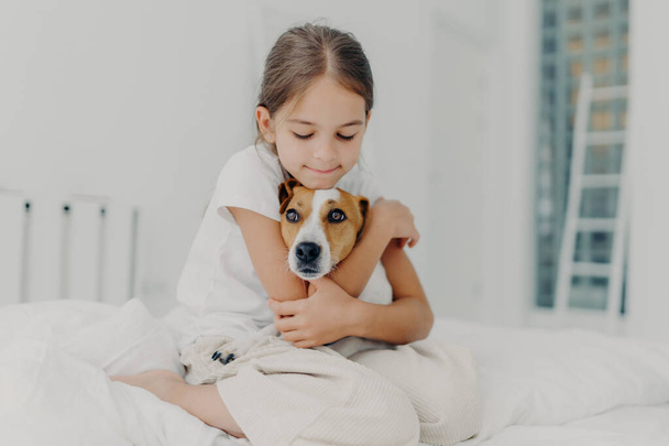Indoor shot of happy small child embraces favourite pet, dressed in pyjamas, expresses love to her dog, plays with animal after awakening, poses on soft bed. Children and domestic animals concept - Photo, Image