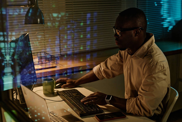 Afrikkalainen amerikkalainen liikemies kirjoittamalla näppäimistön turvakoodit tietokoneella työskennellessään ylitöitä toimistossa - Valokuva, kuva