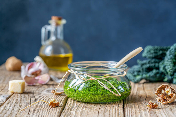 Homemade kale pesto sauce served in a glass bowl over wooden surface and dark background with basic ingredients - kale, walnuts, parmesan, garlic and olive oil. Selective focus. Copy space for text - Foto, immagini