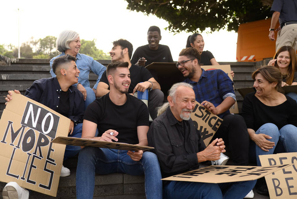 Groep van vrolijke diverse demonstranten van verschillende geslacht en ras zitten op de trap op straat met kartonnen borden en chatten - Foto, afbeelding