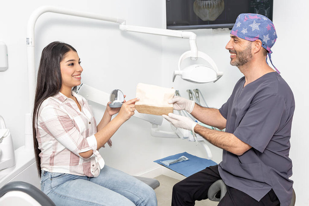 Side näkymä iloinen etninen teini tyttö pitkät tummat hiukset rento vaatteita yhdenmukaistaja laatikko kädessä ottaen reseptiä kädestä hymyilevä mies hammaslääkäri tarkastuksen aikana klinikalla - Valokuva, kuva