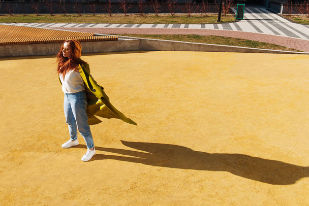 Full length άποψη του τζίντζερ γυναίκα φορώντας κομψό παλτό ποζάρουν στο αστικό στάδιο. Κοκκινομάλλα κοπέλα με γυαλιά ηλίου στέκεται στο δρόμο και απολαμβάνει τον ήλιο - Φωτογραφία, εικόνα