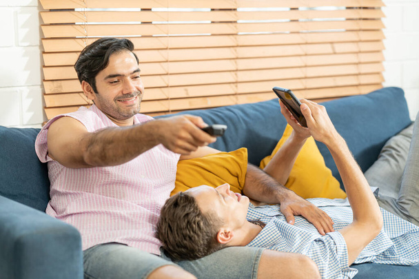 Dos hombres en trajes casuales sentados en el sofá abrazando y viendo la televisión con cerveza y pizza en la sala de estar en casa. Concepto de relación gay LGBT. - Foto, imagen