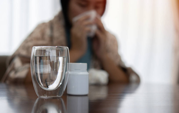 Großaufnahme von Glas Wasser und Medizinflasche mit verschwommener kranker Frau, die unter der Decke auf dem Sofa sitzt und vor Seidenpapier niest. - Foto, Bild