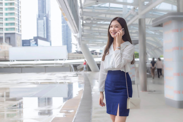 Junge selbstbewusste asiatische Geschäftsfrau geht ins Büro oder an den Arbeitsplatz, die mit einem Smartphone in der Großstadt mit Geschäftshäusern mit der Stadt als Hintergrund. - Foto, Bild