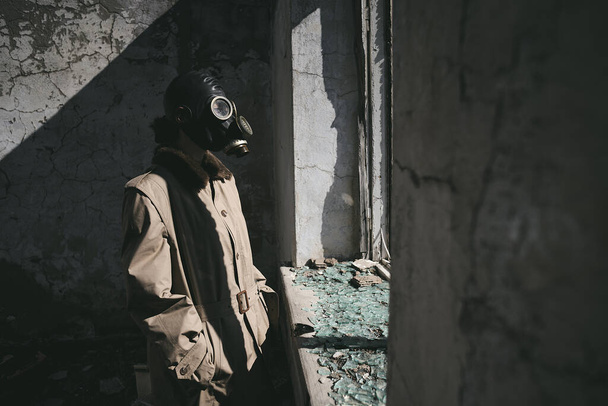a person wearing a gas mask is standing in front of the window of a destroyed house, good for book cover - Фото, изображение