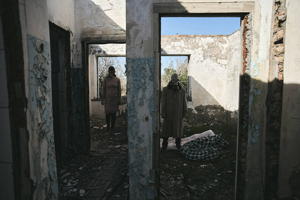 Ένας άνδρας και μια γυναίκα σε ένα κατεστραμμένο κτίριο, με μάσκες αερίου, στέκονται σε διαφορετικά δωμάτια, καλό για το εξώφυλλο του βιβλίου - Φωτογραφία, εικόνα