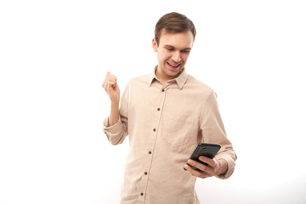 Portrait of young handsome caucasian man using mobile phone and smiling, clenching fist celebrating victory isolated on white studio background, joyful facial expression - Photo, image