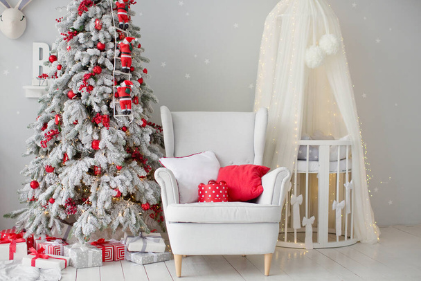 Christmas tree with red decorations, white armchair with cushions and baby bed crib with canopy in cute childs room interior with grey walls - Photo, Image