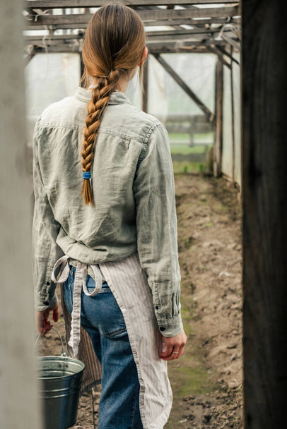 Una mujer atractiva sosteniendo un cubo de lata y caminando en el invernadero mirando las plántulas  - Foto, imagen